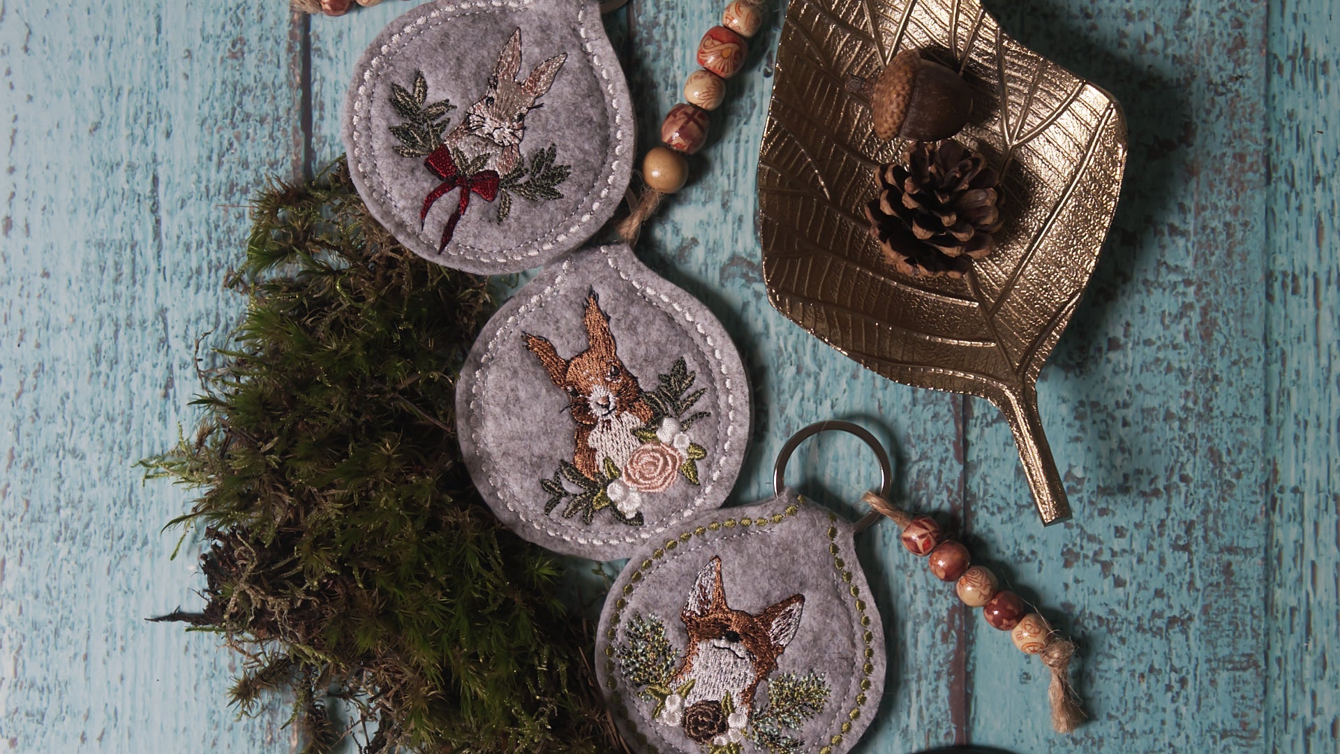 Gestickte ITH Schlüsselanhänger aus grauem Filz mit liebevollen Tiermotiven – Hase und Eichhörnchen mit floralen Details, dekoriert mit Moos, Holzperlen und goldener Blattschale auf rustikalem Holzhintergrund.