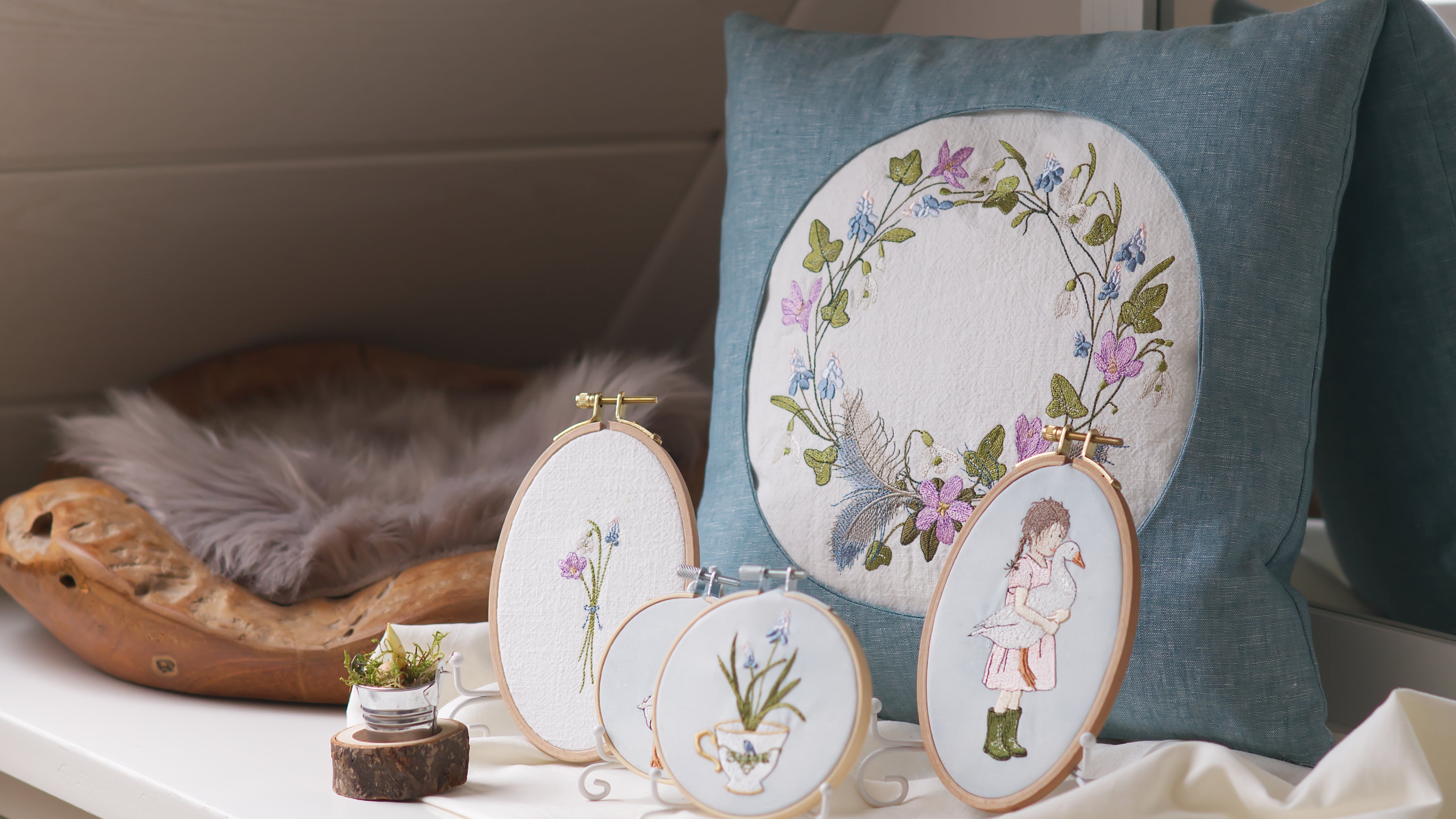 Kissen und Stickrahmendekorationen mit Bestickung von Frühlingsmotiven, wie Kranz, Mädchen mit Gans, Schneeglöckchen in der Teetasse und Frühblüherstrauss